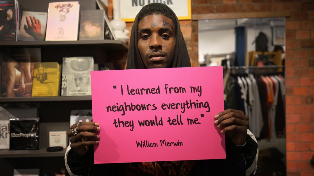 Man in a clothes store holding a quote by William Merwin reading: "I learned from my neighbours everything they would tell me."