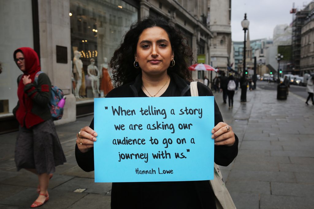 Woman standing on a street holding up a quote to camera reading: "When telling a story we are asking our audience to go on a journey with us", quote by Hannah Lowe 