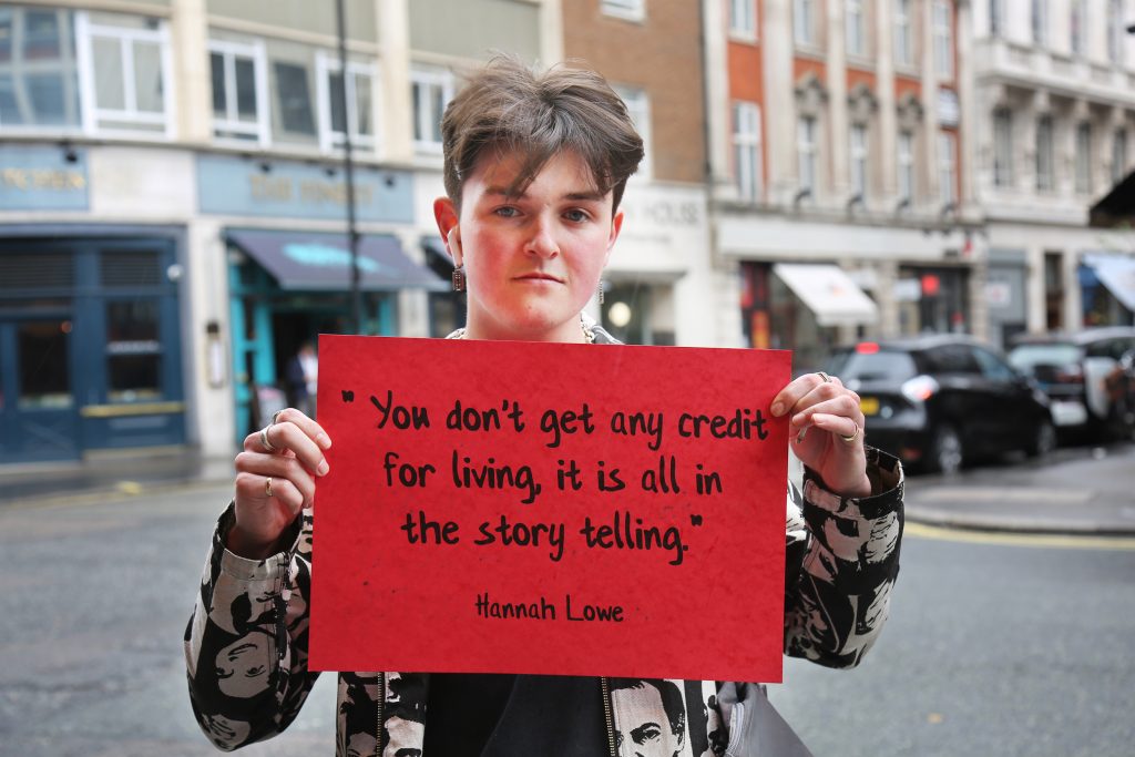 person on the street holding a card with a quote on it "You don't get any credit for living, it is in the story telling" , quote by Hannah Lowe 